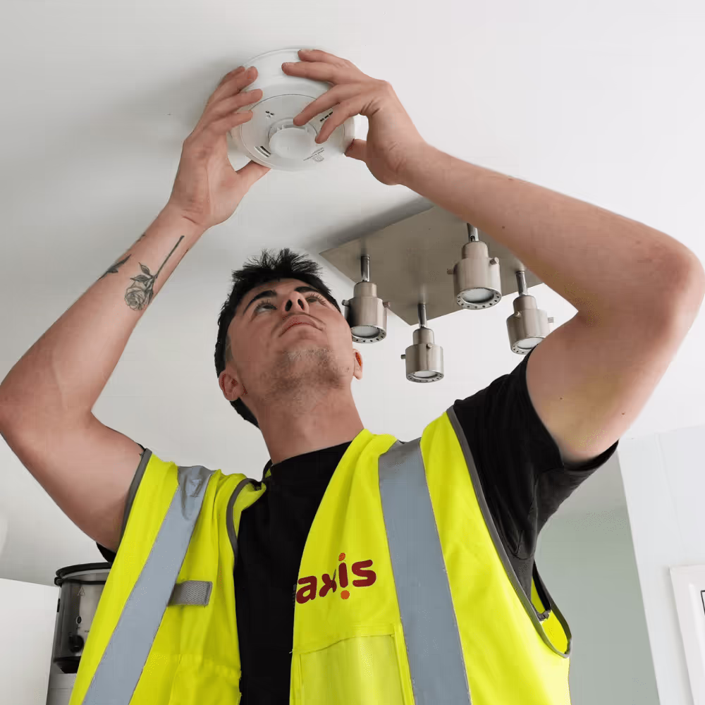 A male Axis technician in a high-visibility vest is installing a smoke detector on a white ceiling, demonstrating fire safety and maintenance work often paired with energy upgrades.